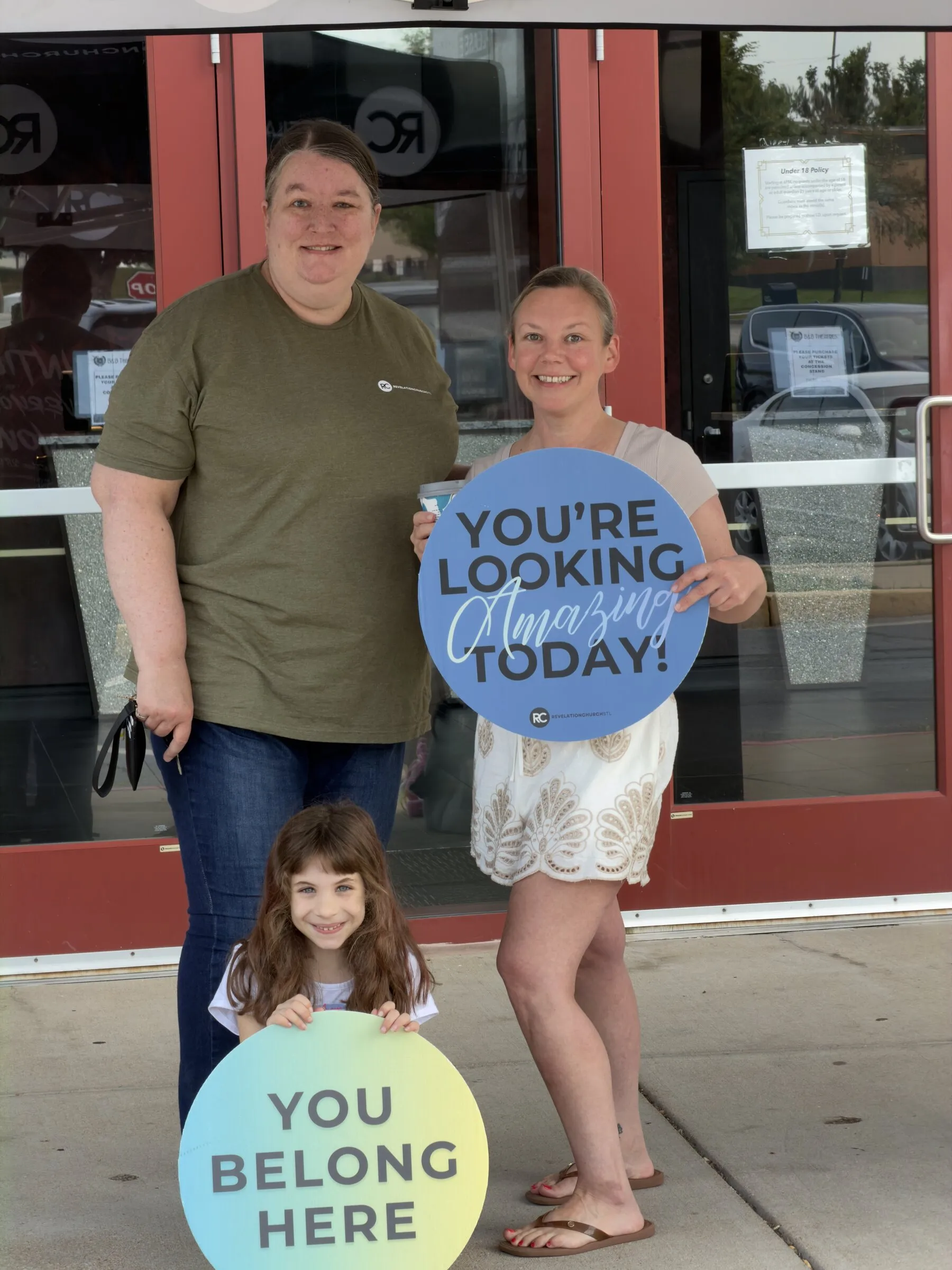 Two welcome-team members and a child holding “You belong here” and “You’re looking amazing today” signs outside the theater